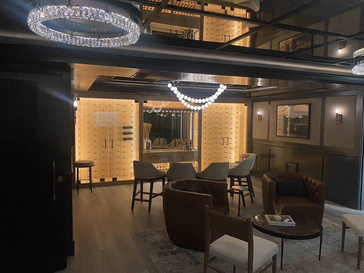 A dark, paneled lounge with a crystal ring chandelier, a strand of globe lights and a backlit wine wall behind a bar.