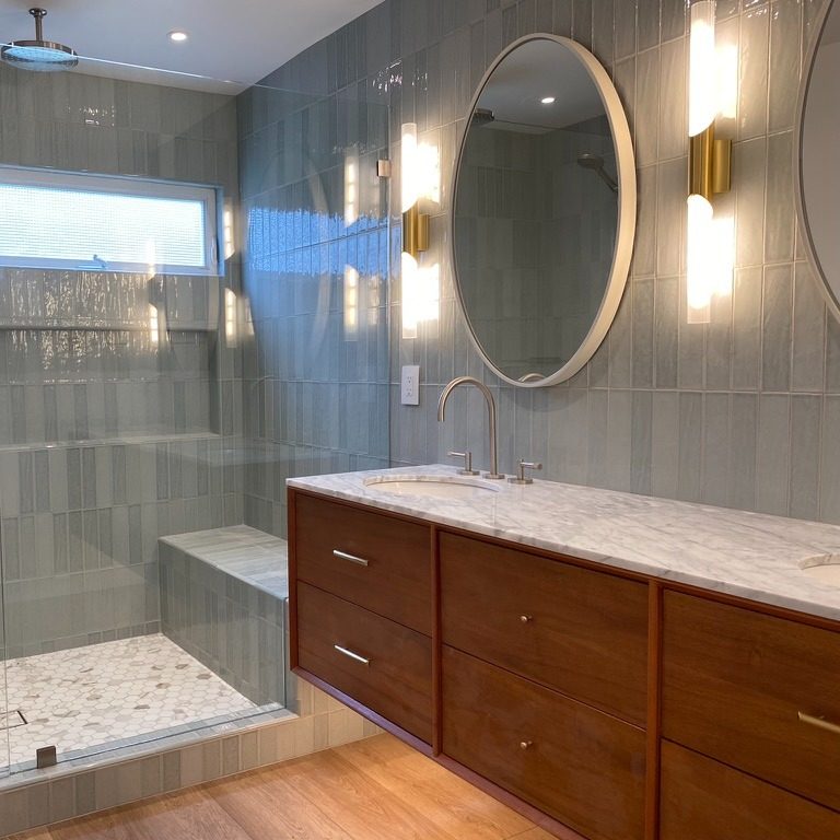 A tiled bathroom with brass tube sconces on either side of a round mirror over a wood vanity.