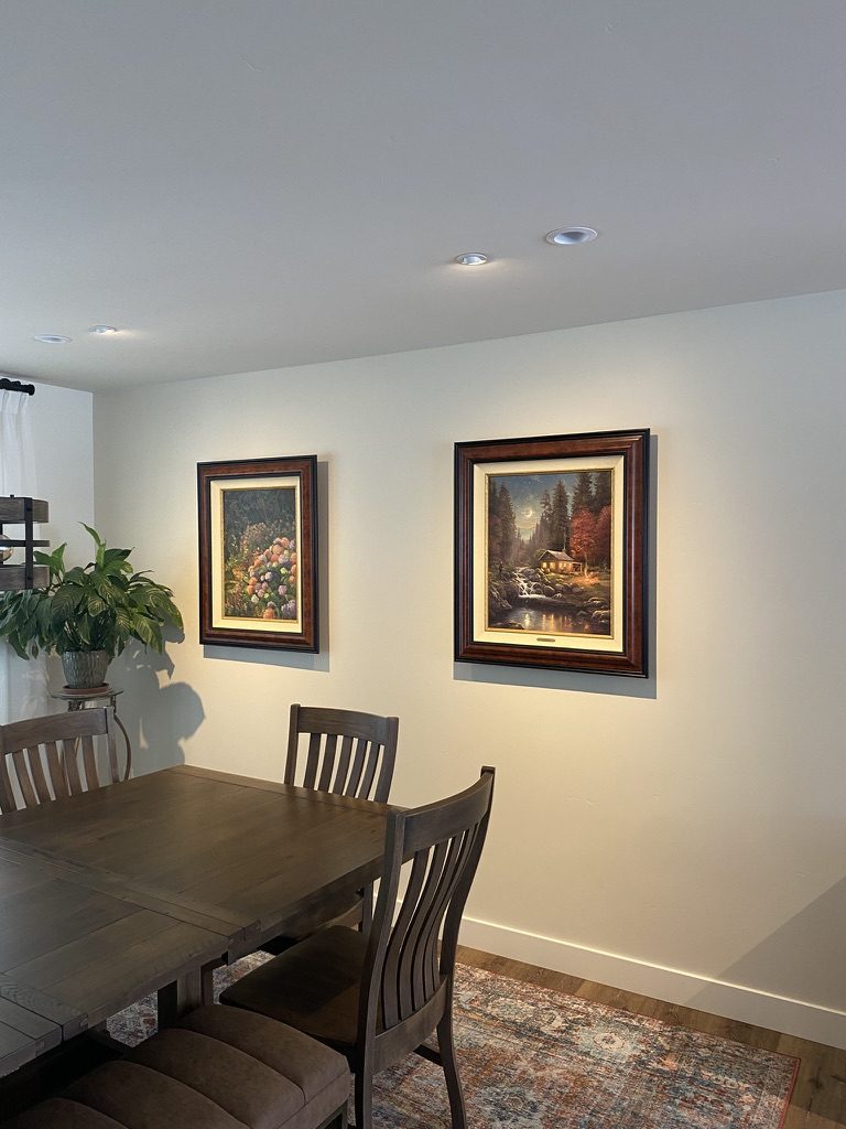 A dining room with recessed ceiling lights aimed at two framed paintings above the table.