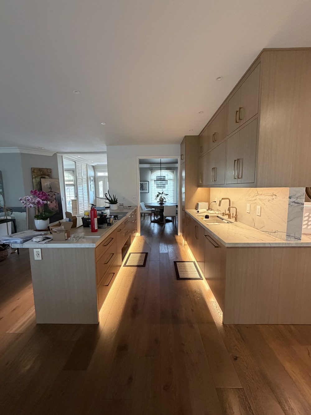 A long kitchen with pale wood cabinets, marble counters and a warm strip of light glowing along the floor under the cabinets and the island.