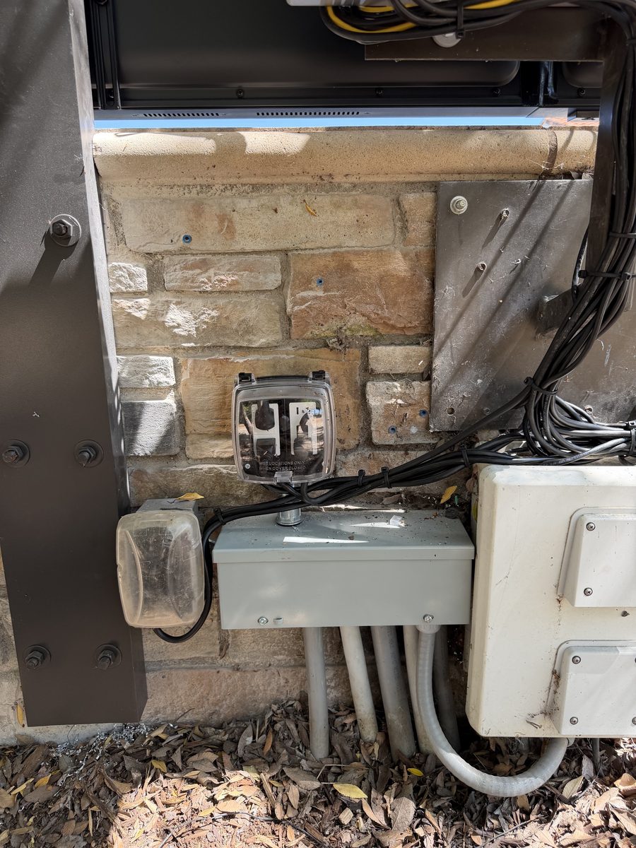Outdoor electrical equipment against a stone wall: a gray box, a covered outlet and conduit running into the ground.